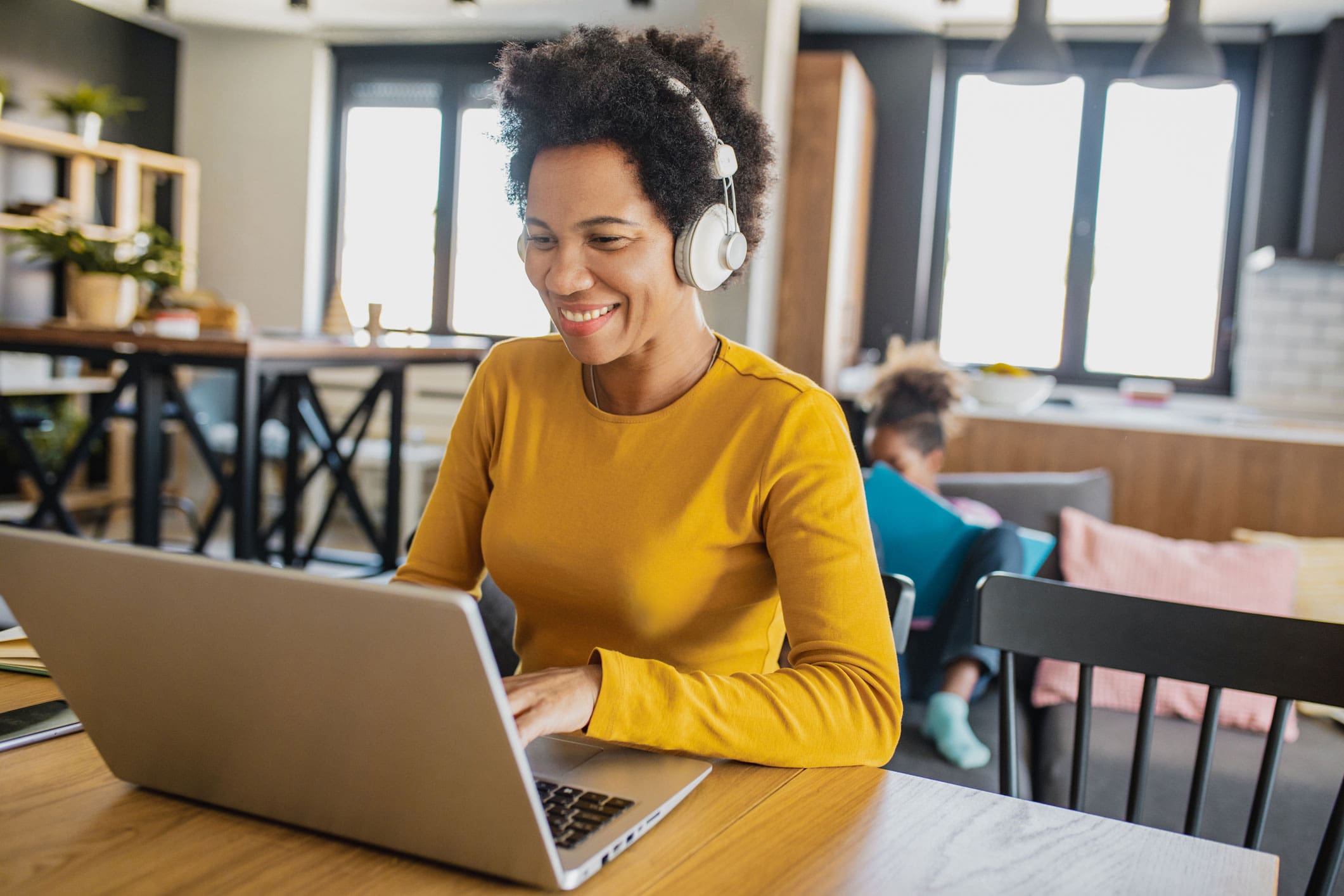 A woman in a gold shirt participates in an online class while children play in the background.